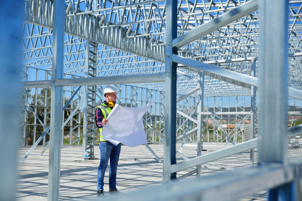 Electrician on construction site reviewing blueprints amid steel framing, representing project management and timely conduit and fittings delivery.